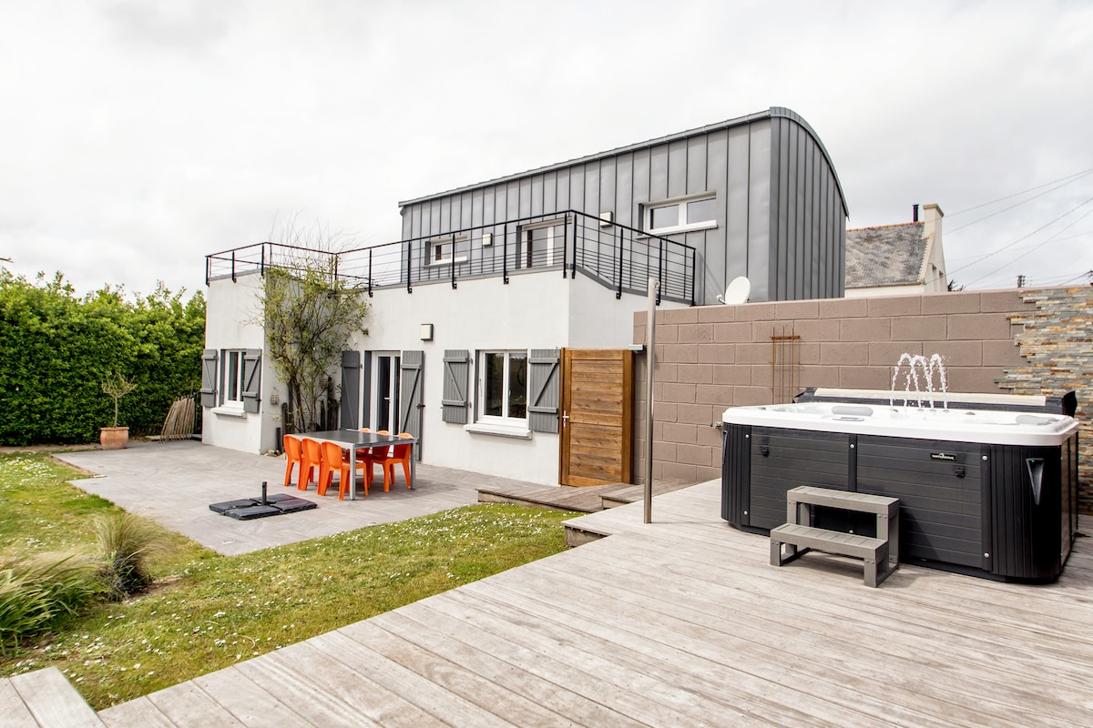 The exterior of the house features a spacious deck leading to a private garden. A six-person hot tub is positioned alongside a seating area with an orange dining table and chairs. The building showcases modern architecture with a combination of dark and light elements.