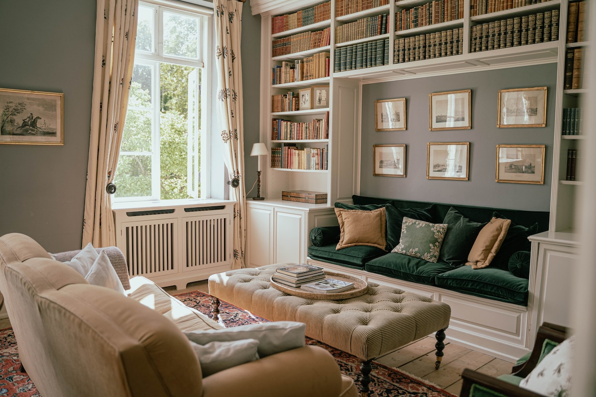 A stylish living area features a comfortable beige sofa and a plush green bench, accompanied by a coffee table stacked with books. Decorative framed art adorns the walls, while a bright window allows natural light to illuminate the space, enhancing the cozy ambience.