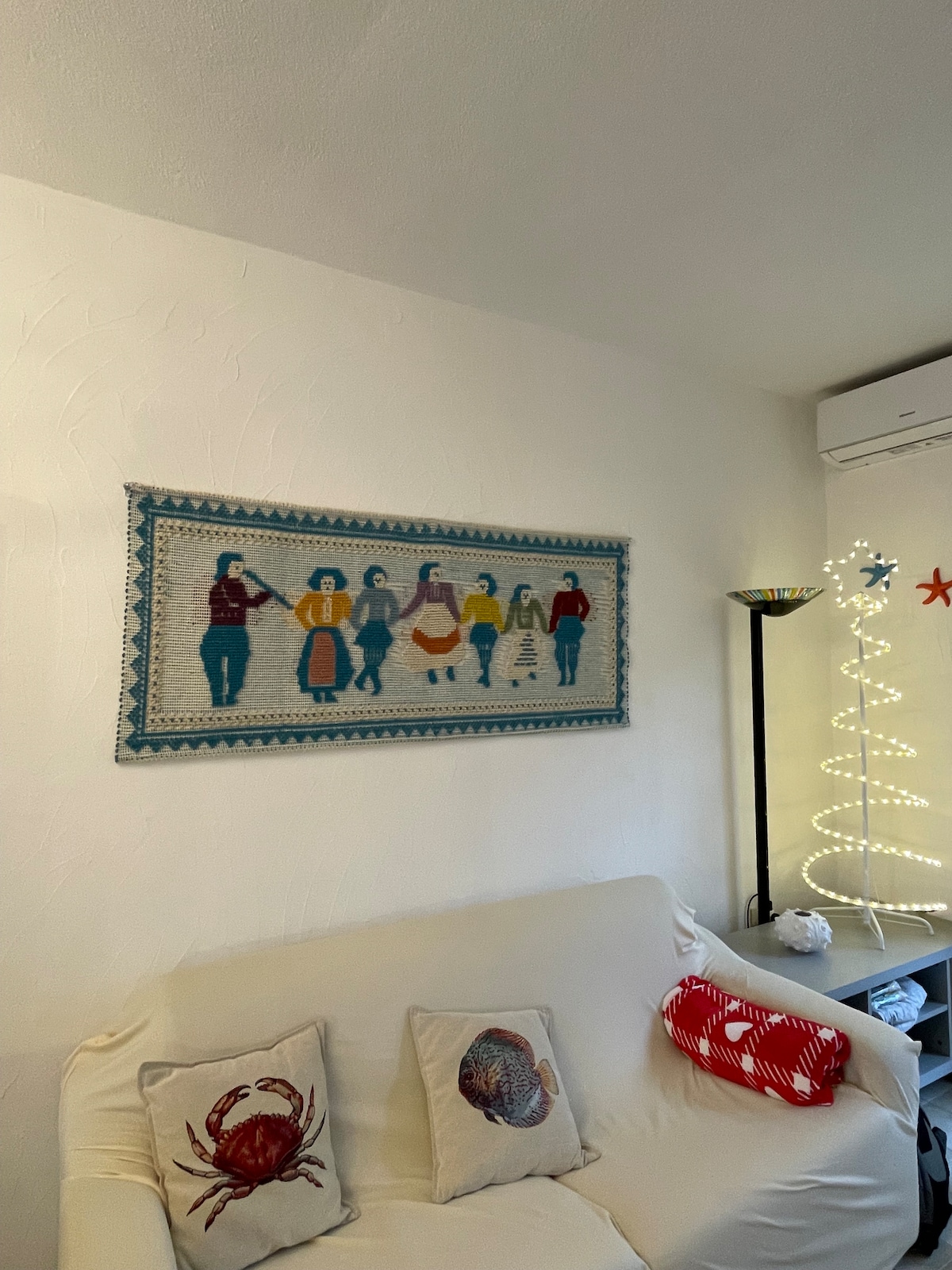 A wall tapestry depicting figures in traditional attire is showcased above a light-coloured sofa with two decorative cushions featuring sea-themed designs. A slim lamp with a colorful shade stands next to the sofa, illuminated by soft lighting. A white, minimalist Christmas tree is adorned with lights in the background.