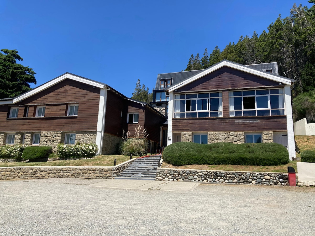 Two charming buildings are situated side by side, featuring a combination of wooden and stone exteriors. Lush greenery surrounds the entrance, and a clear pathway leads to the doorsteps. The buildings are set against a backdrop of tall trees under a clear blue sky.