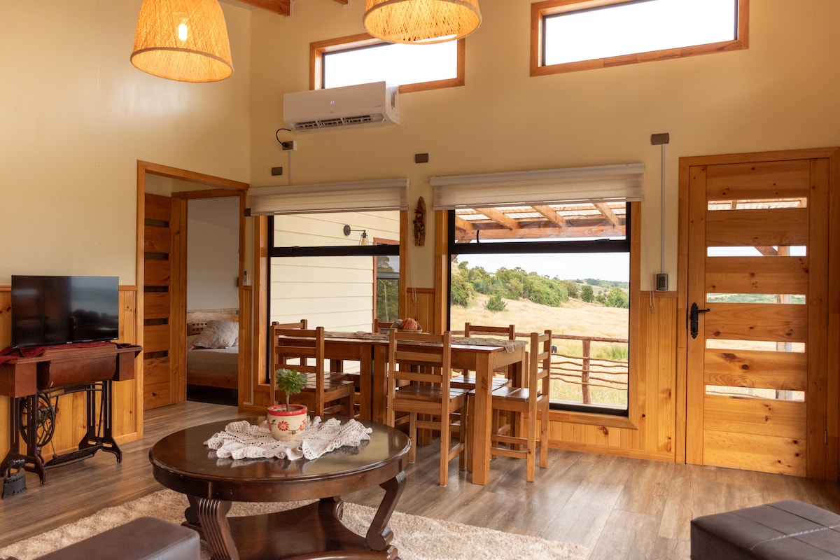A welcoming interior space is highlighted, featuring a dining table surrounded by wooden chairs. Natural light fills the area through large windows, and a comfortable seating arrangement is positioned near a TV. Wooden accents and light-colored walls enhance the cozy atmosphere.