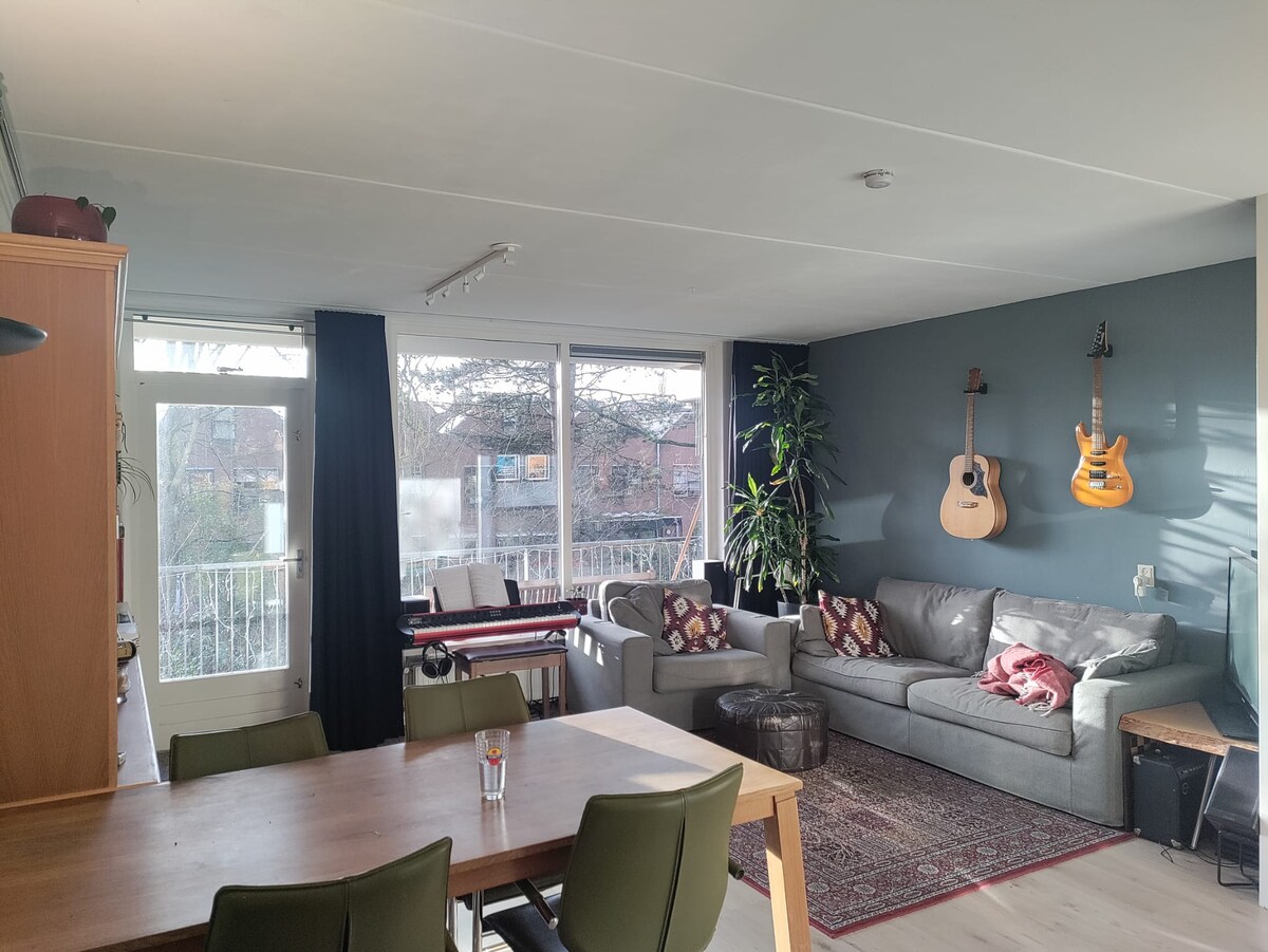 A spacious living area features a comfortable gray sofa and two acoustic guitars mounted on the wall. A dining table with several chairs is positioned in the foreground, while large windows allow natural light to fill the room, enhancing the welcoming ambiance.