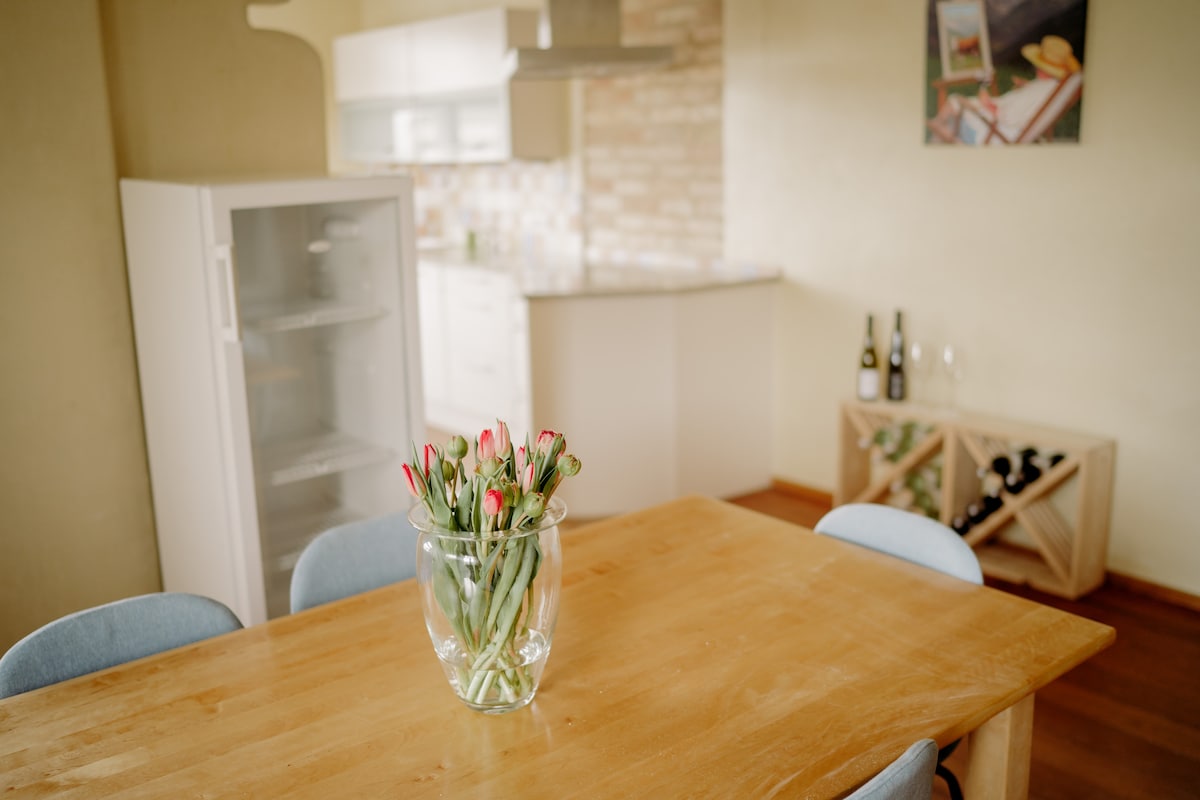 A light-filled dining area features a wooden table surrounded by soft blue chairs. A vase filled with fresh flowers sits at the center, adding a touch of color. In the background, a modern kitchen with a stylish backsplash and essential appliances is visible.