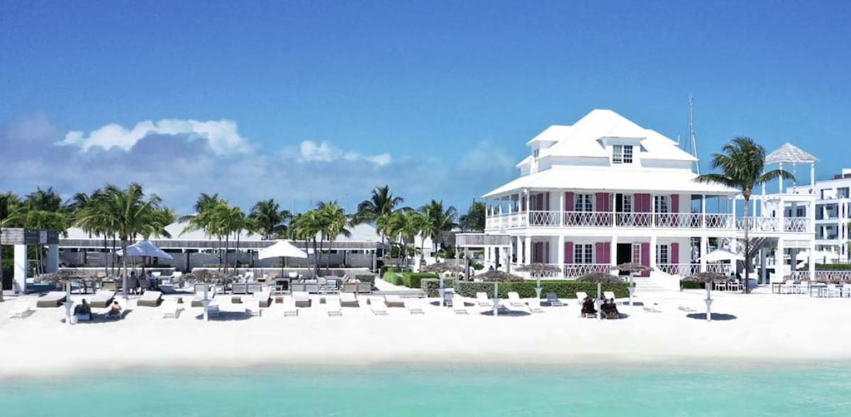 A classic coastal building is situated against a clear blue sky, featuring multiple balconies and large windows. The foreground displays a sandy beach lined with comfortable lounge chairs and shaded areas, surrounded by palm trees, creating a spacious outdoor relaxation area.