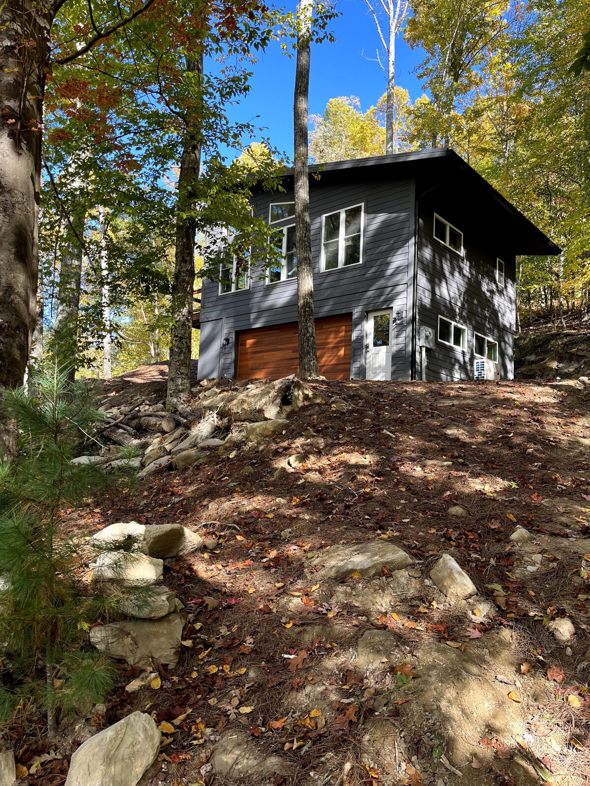A modern home is nestled among the trees on a sloped lot, showcasing a striking dark exterior with large windows. Natural rock formations and fallen leaves surround the building, contributing to a serene woodland setting under a clear blue sky.