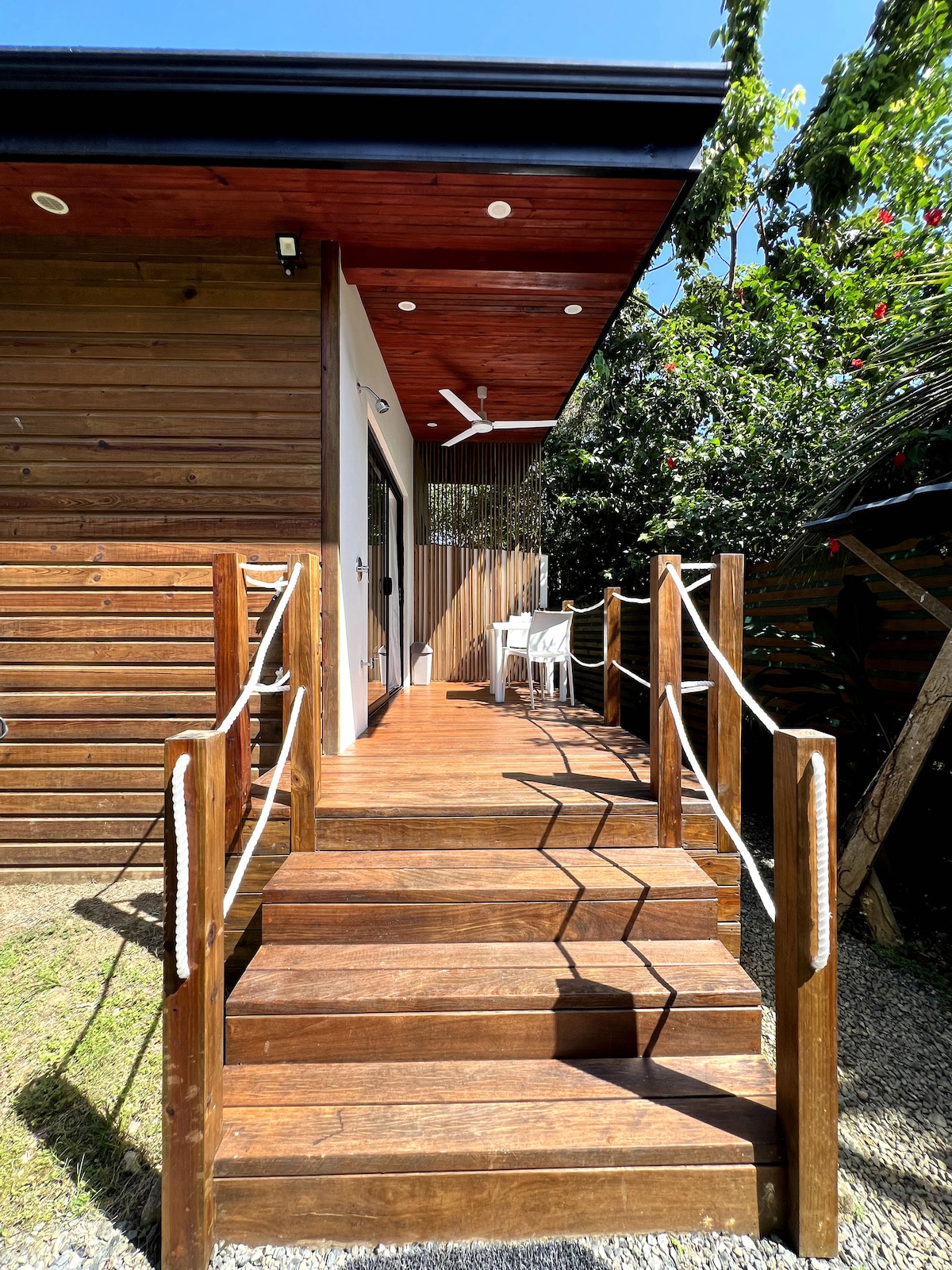 A wooden staircase leads to the entrance of the studio, surrounded by lush greenery. The steps are framed with rope railings, providing an inviting approach to the space. A chair can be seen on the private terrace, enhancing the outdoor setting.