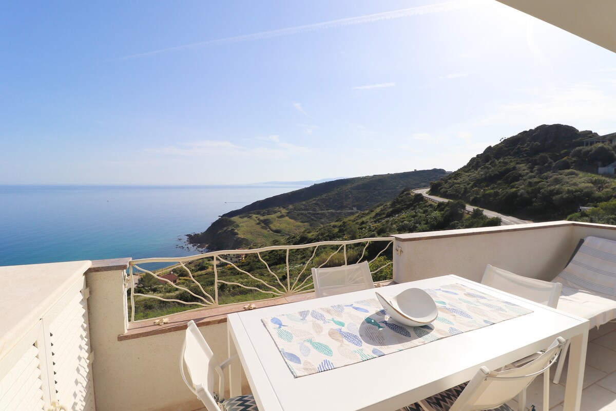 A spacious outdoor terrace features a large dining table set with a patterned table runner, overlooking a panoramic view of the sea and lush green hills. White outdoor chairs complement the serene landscape, creating an inviting space for meals and relaxation.