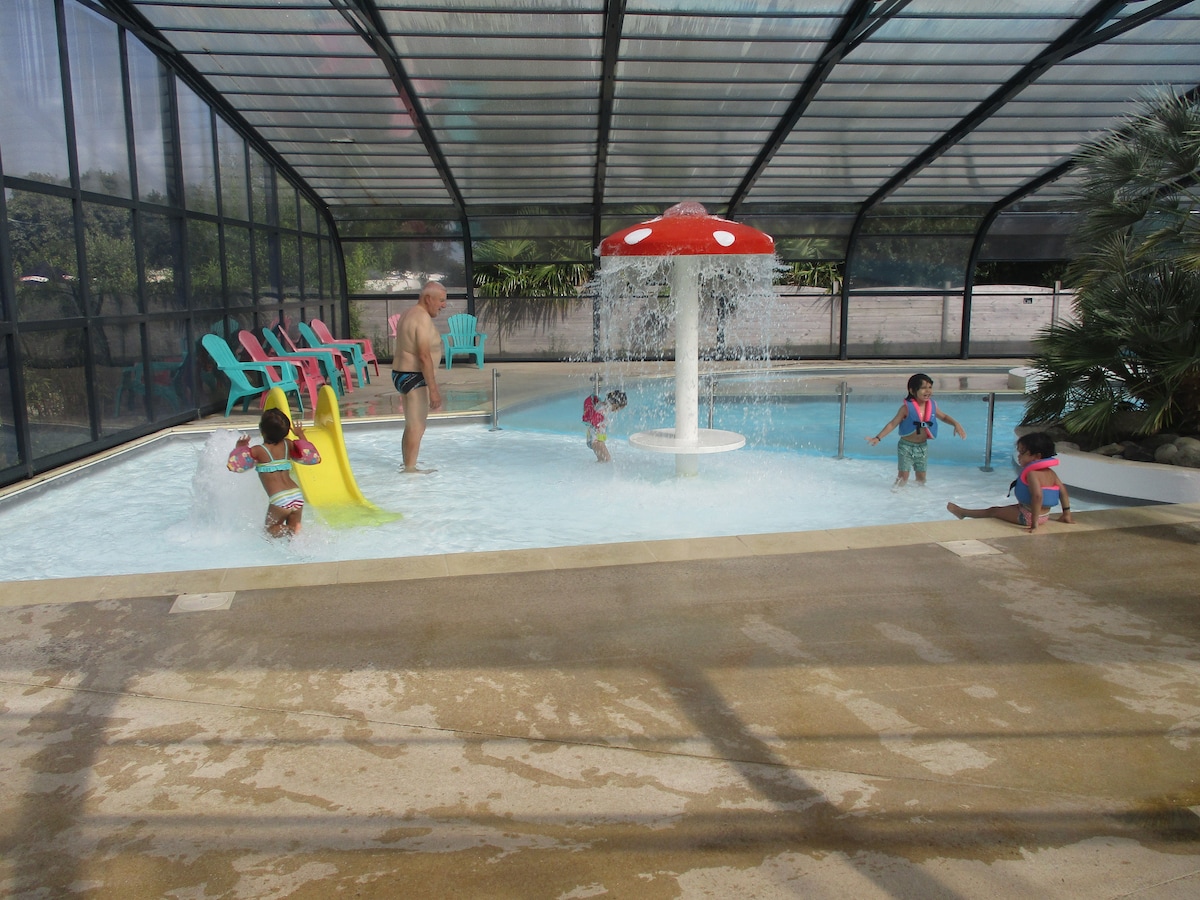An indoor pool area is depicted, featuring a vibrant red mushroom water feature. Colorful lounge chairs line one side, while several children enjoy the shallow water, including one sliding down a yellow slide. Sunlight filters through the transparent ceiling, creating a bright atmosphere.