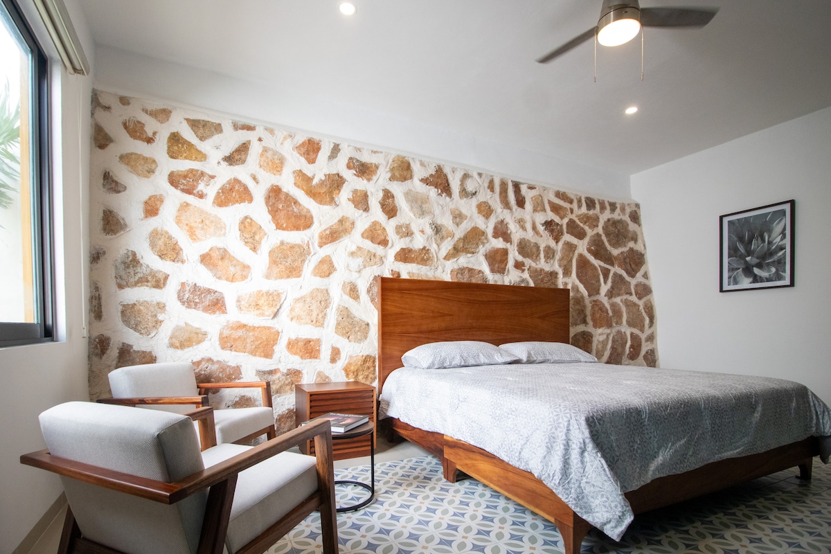 A spacious bedroom features a natural stone accent wall, adding texture to the modern design. A comfortable bed is positioned alongside stylish armchairs, offering a cozy seating area. Natural light is provided by a window, while a ceiling fan enhances air circulation in the room.