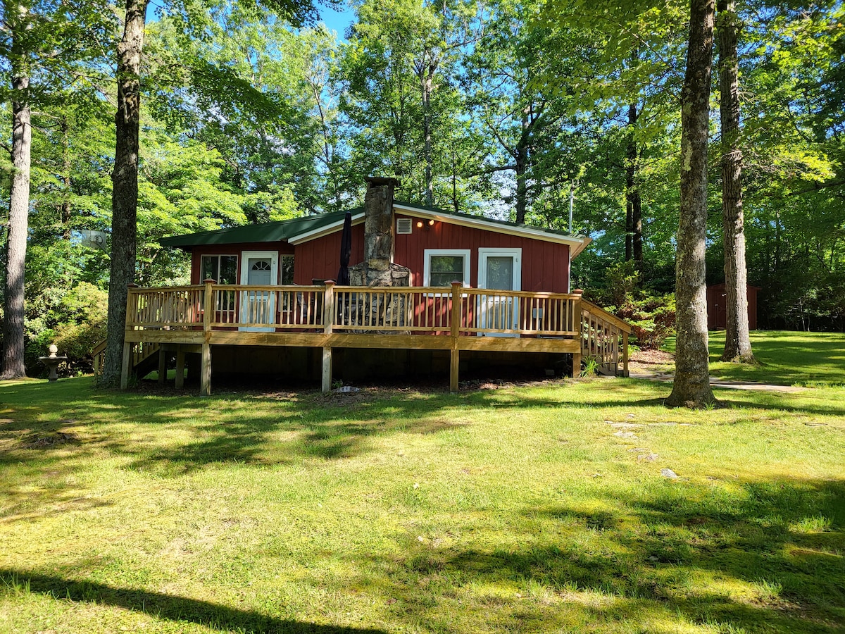 A secluded cottage is surrounded by lush greenery, featuring a wooden deck that wraps around the front. Large windows offer views of the exterior landscape. A stone chimney is visible, suggesting a cozy fireplace within, and the grassy area is dappled with sunlight filtering through the trees.