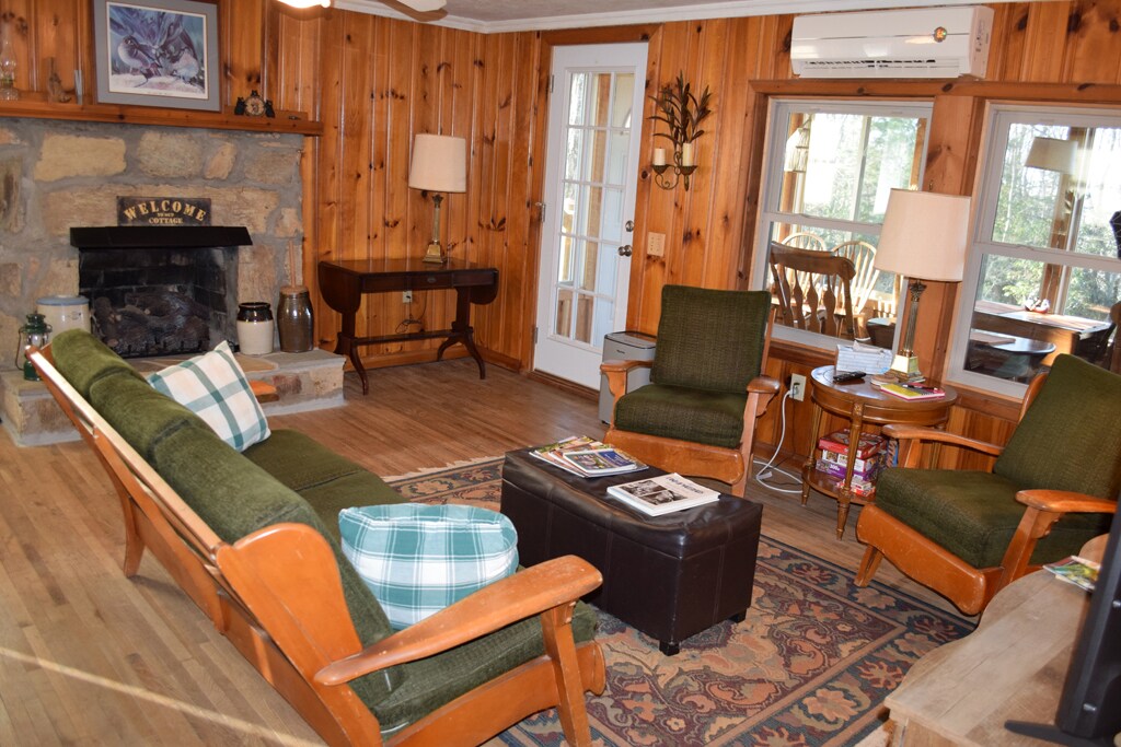 A rustic living room features natural wood paneling and a stone fireplace. Comfortable seating includes two green upholstered armchairs and a wooden sofa with decorative pillows. A coffee table with magazines sits centrally, complemented by a side table and a glass door leading outside.