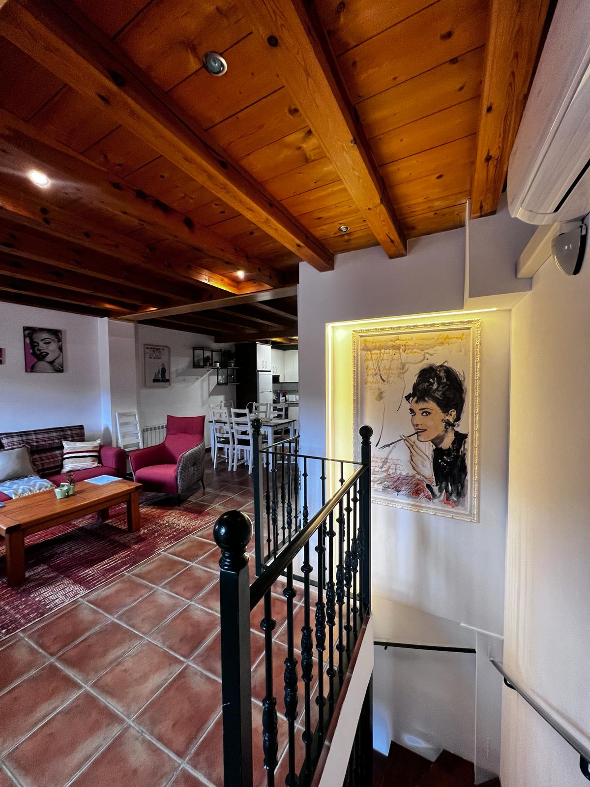A well-lit staircase leads to an upper level, framed by black iron railings. The airy space features wooden beams and terracotta tiles. A cozy sitting area with a wooden coffee table and assorted seating is visible, along with artwork adorning the walls.