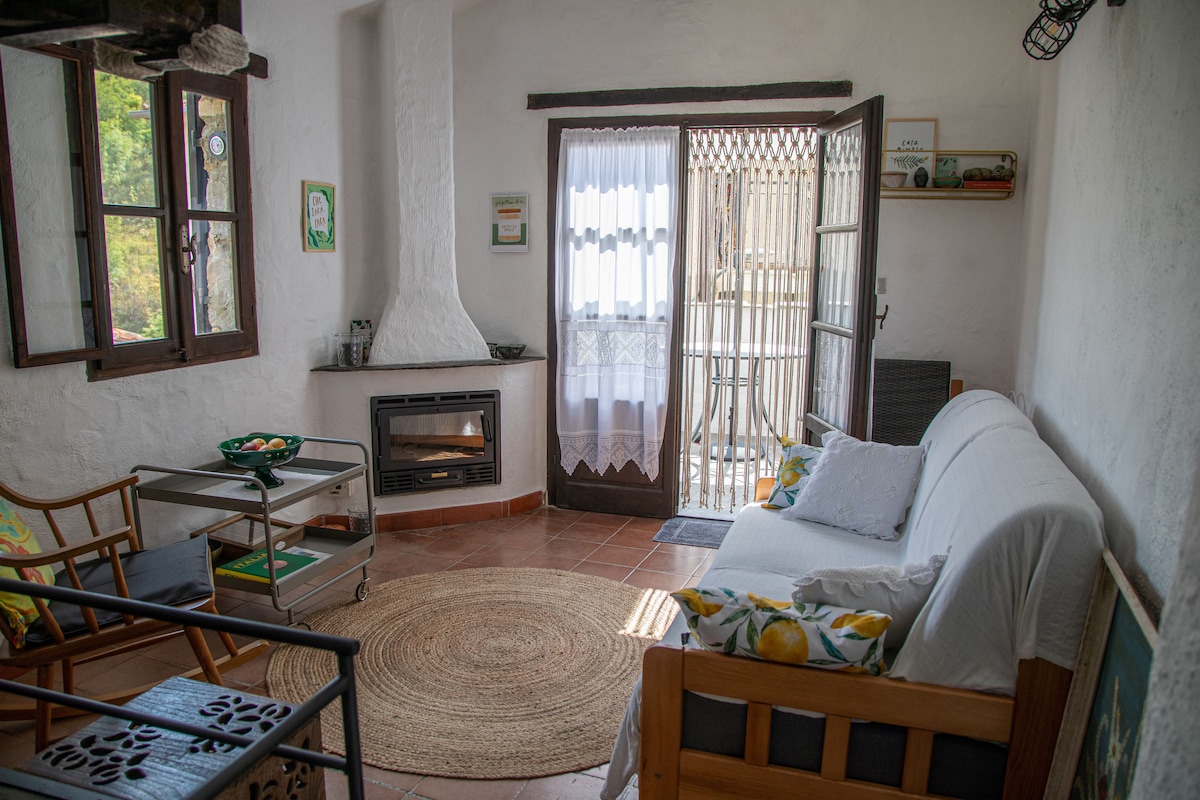 A small living area includes a comfortable couch and a rocking chair, with a rug on the tiled floor. Natural light enters through the double doors leading to the terrace, complemented by a fireplace and decorative wall art.