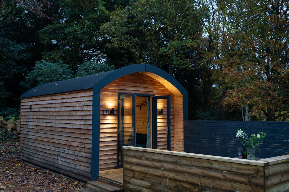 A timber-framed glamping pod is surrounded by trees, featuring an open deck with wooden railings. The entryway is highlighted by large glass doors, inviting natural light, while soft lights on the walls create a warm ambiance in the evening.