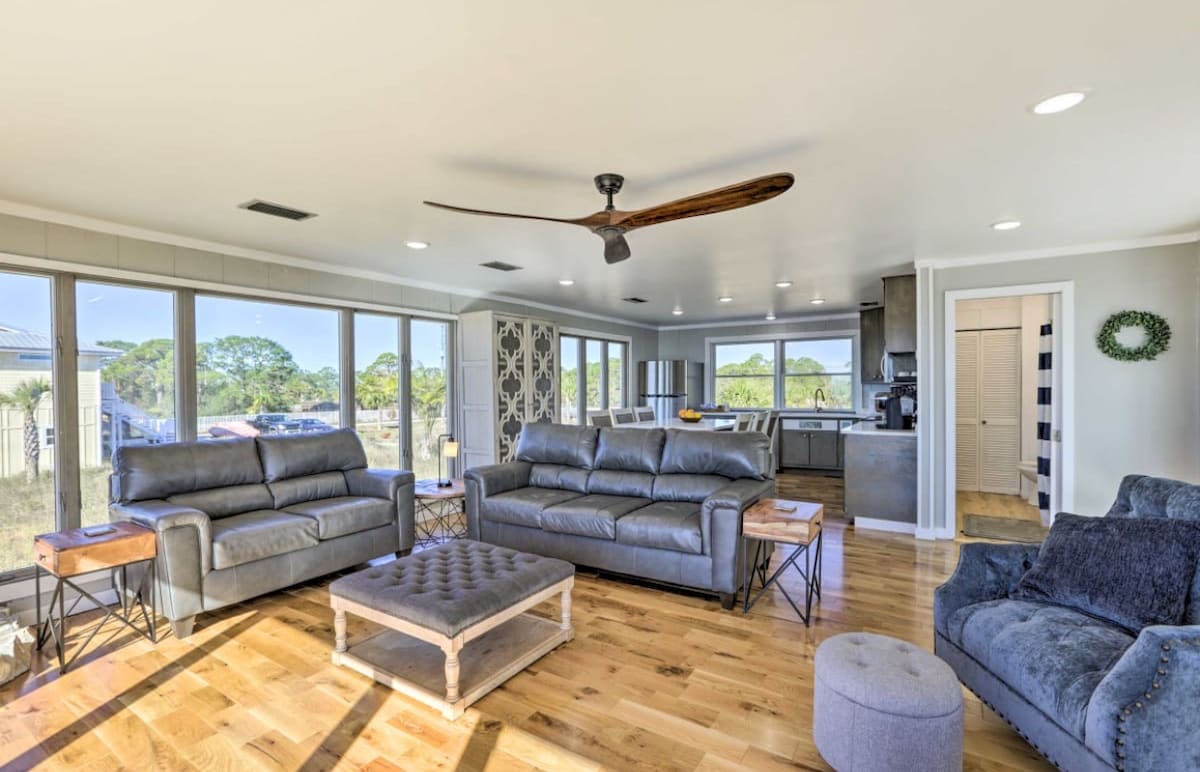 A spacious living area is highlighted by large windows that offer ample natural light and views of the surrounding landscape. Modern furnishings include two gray sofas and a gray armchair, complemented by a wooden coffee table and side tables. The open layout connects seamlessly to the kitchen.