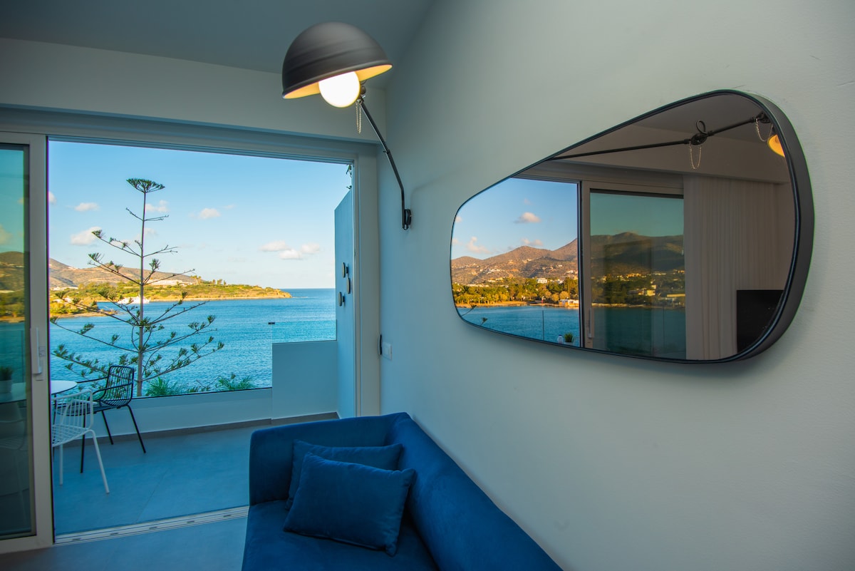A bright living area is furnished with a blue sofa and features a large mirror on the wall. A sliding door opens to a balcony, providing a view of the Mirabello Bay and surrounding hills. Sunlight highlights the inviting coastal scenery.