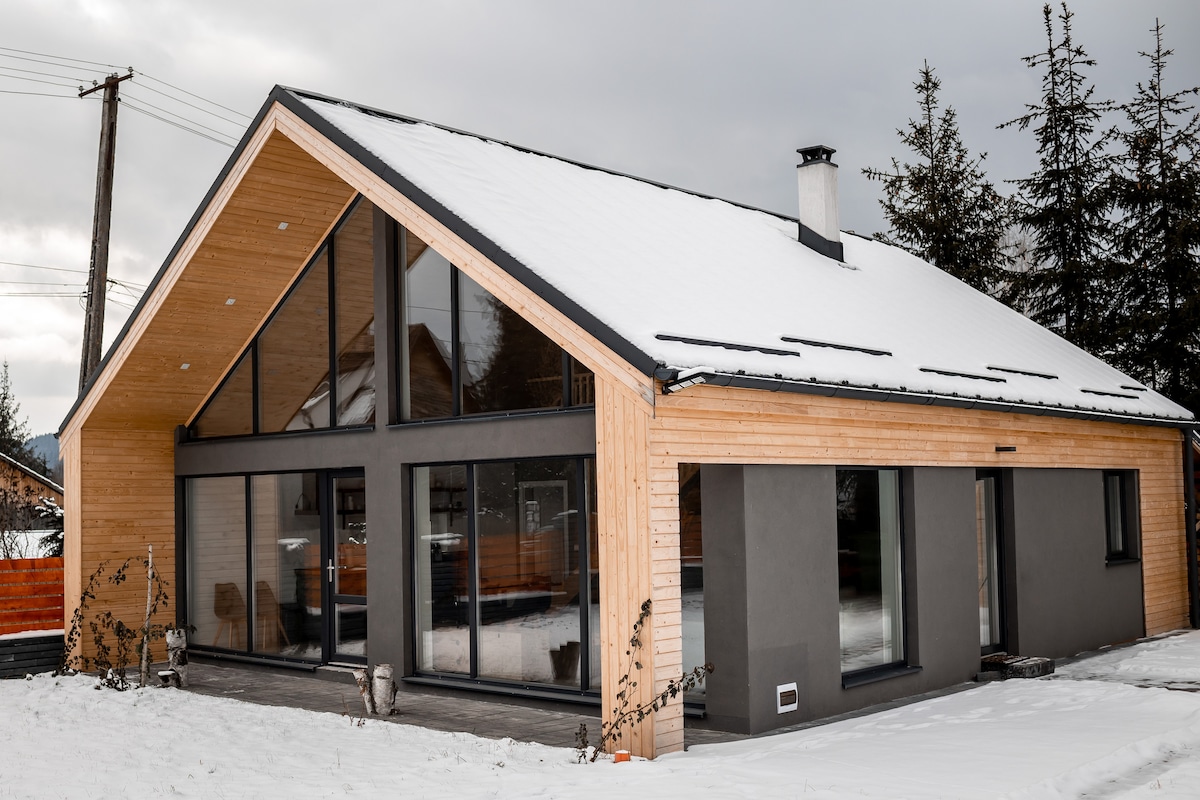 A modern home features a combination of wood and stone materials, with large glass windows that highlight the exterior design. Snow covers the ground, creating a serene atmosphere. A sloped roof with clean lines and tall pine trees frame the structure.