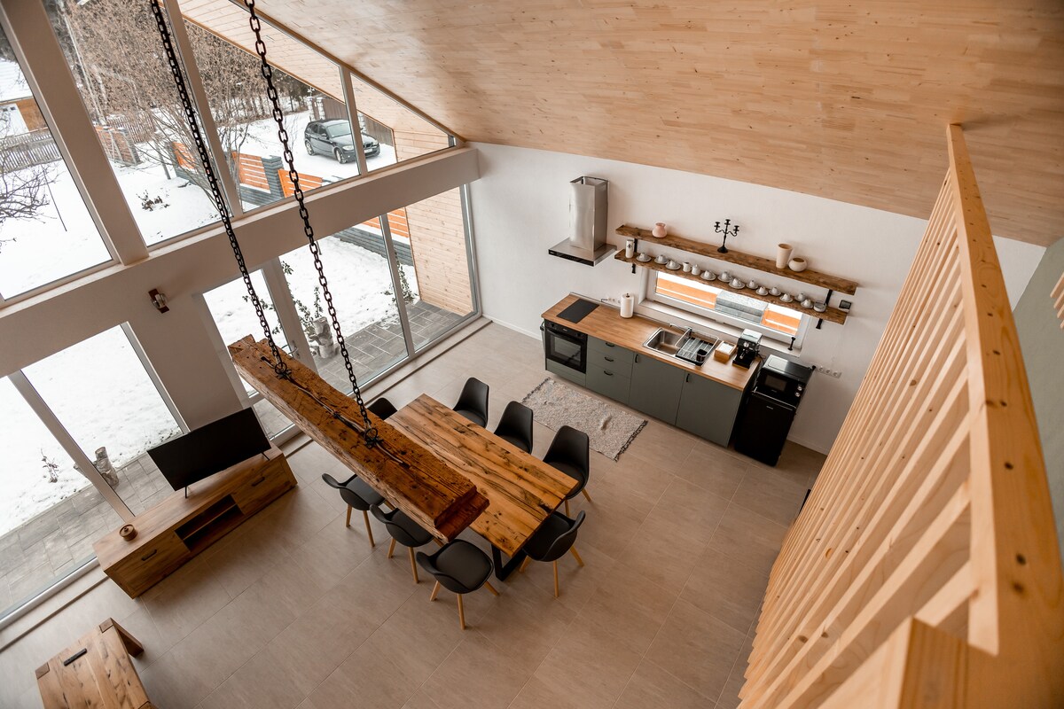 An open-concept living space features a high, wooden ceiling with large windows allowing natural light. A long, wooden dining table is surrounded by black chairs. The kitchen area is equipped with modern appliances and a wooden shelf above the countertop.