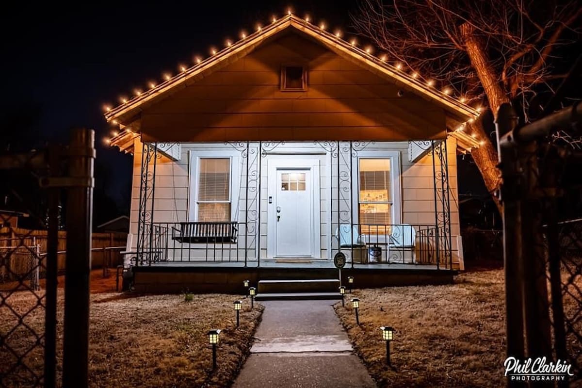 The exterior of the house is illuminated at night with warm string lights along the roofline. The porch features a bench and two lit windows, creating a welcoming entrance. A pathway lined with solar lights leads to the front door, enhancing the inviting atmosphere.