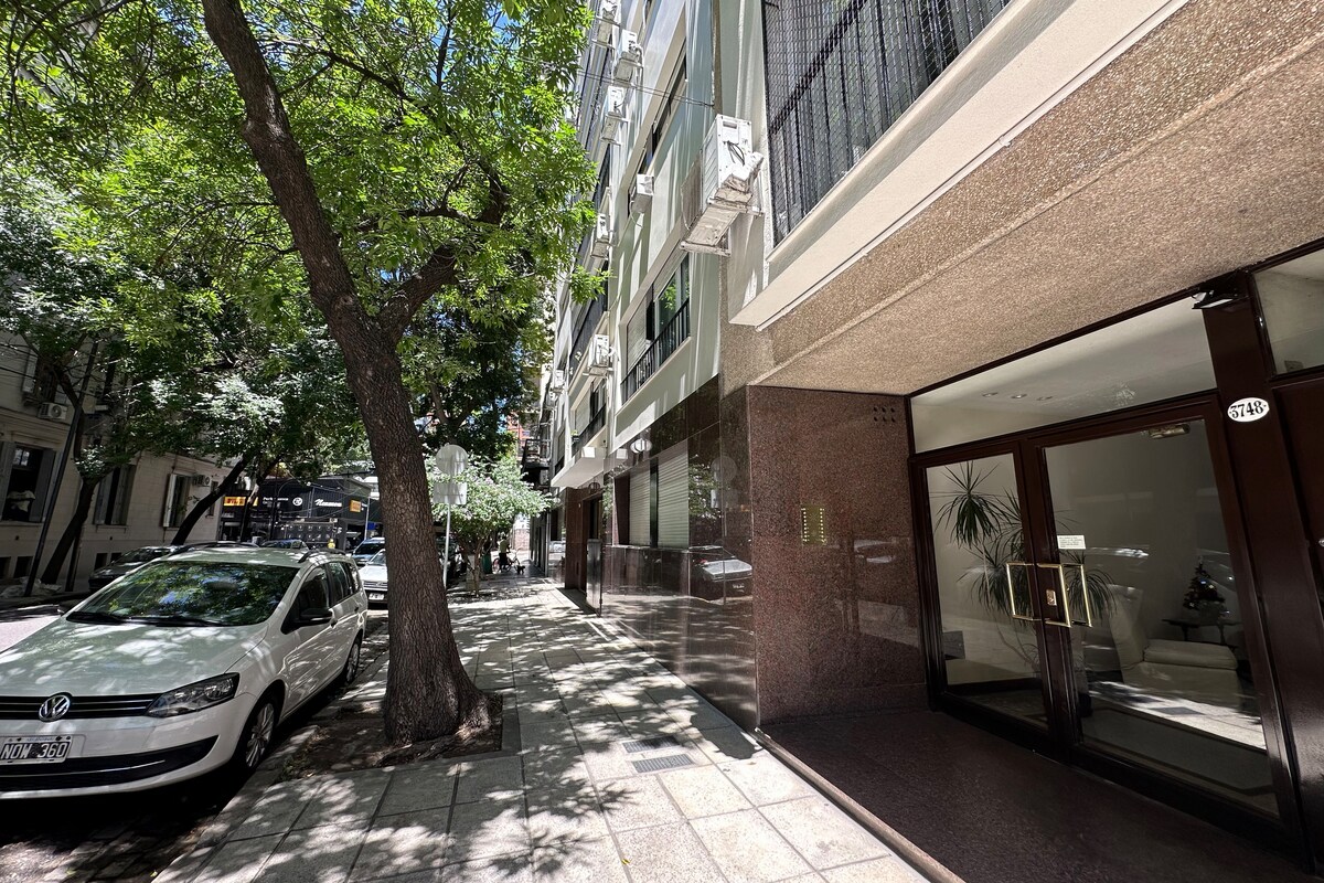 A tree-lined street is featured, with sunlight filtering through the branches. The entrance of a modern building is visible, showcasing polished surfaces and large glass doors. Parked vehicles can be seen on the side of the street, contributing to the urban atmosphere.