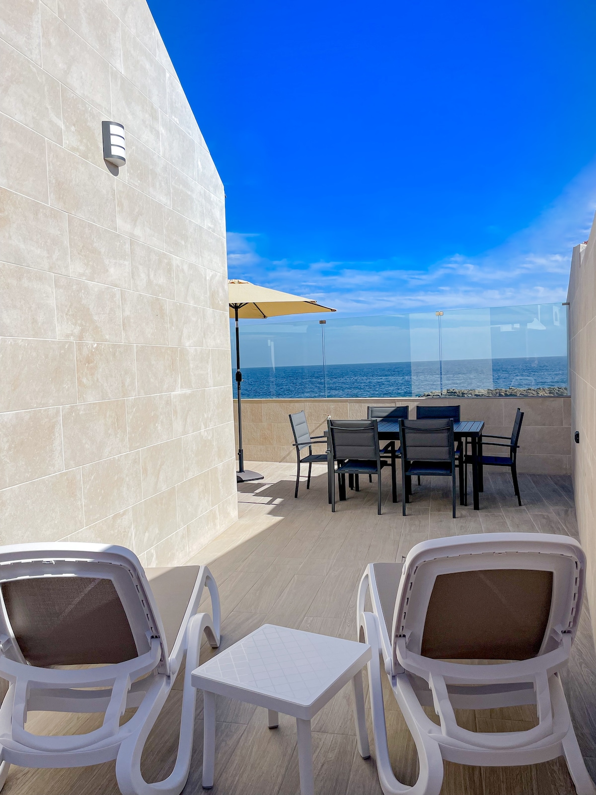 An outdoor terrace features two white lounge chairs and a small side table, with a dining set for six under a large umbrella. The sea is visible beyond a glass barrier, complemented by a clear blue sky.