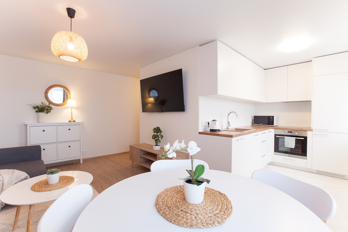 A modern living area features a light-colored kitchen with built-in appliances, including an oven and microwave. A large flat-screen TV is mounted on the wall, opposite a cozy couch. A round dining table with a woven centerpiece and a small potted plant adds a touch of greenery.