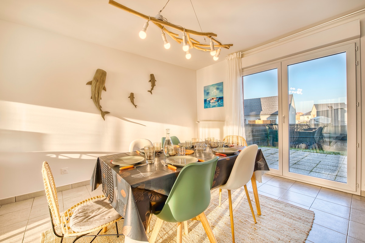 A dining area is illuminated by soft lighting from a unique fixture above a large table set for a meal. Comfortable chairs surround the table, while large glass doors lead to an outdoor space, providing a view of the surroundings and allowing natural light to fill the room.