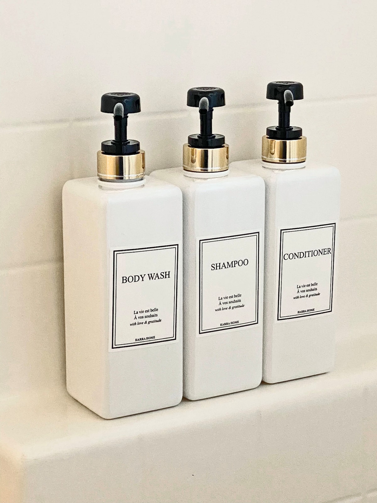 Three neatly arranged, minimalist pump bottles are displayed in a bathroom setting. The labeled containers contain body wash, shampoo, and conditioner, each featuring elegant black and white design elements against a light tile background.