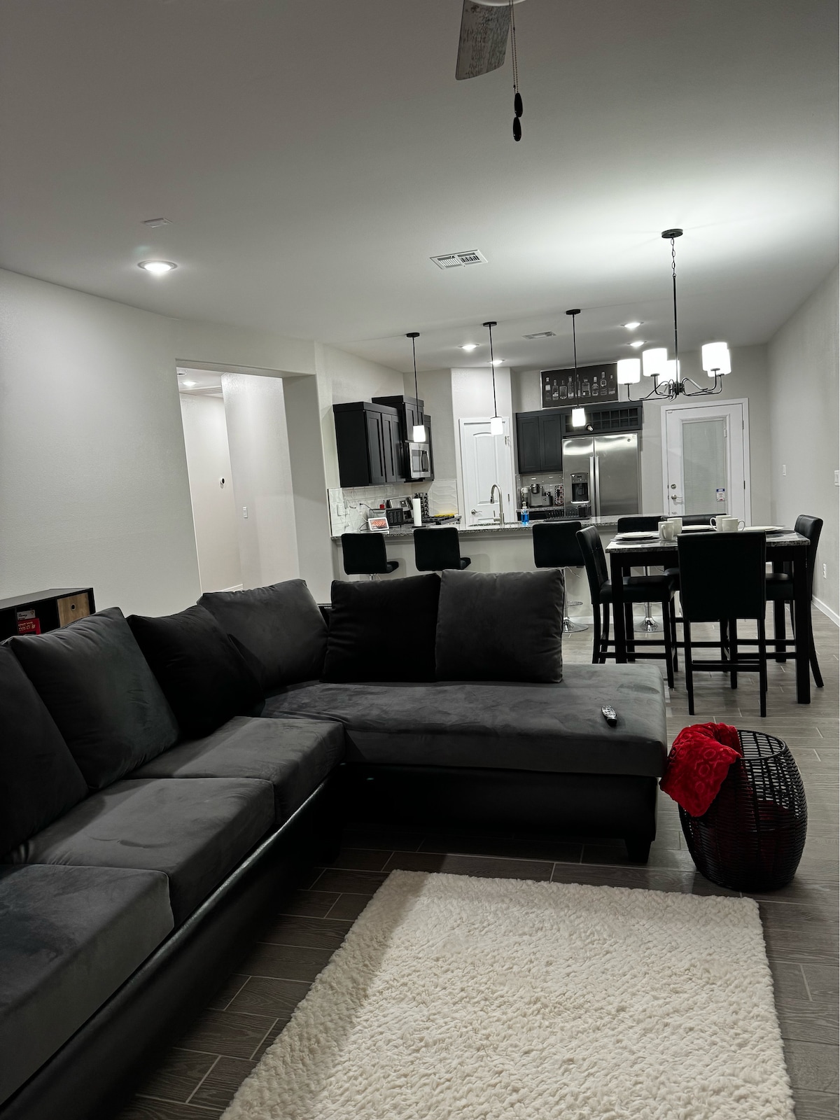 A modern living area features a dark sectional sofa with multiple cushions. A light-colored rug lies on the tiled floor, complementing the sleek furniture. The kitchen and dining space are visible in the background, equipped with stainless steel appliances and a dining table with high-backed chairs.