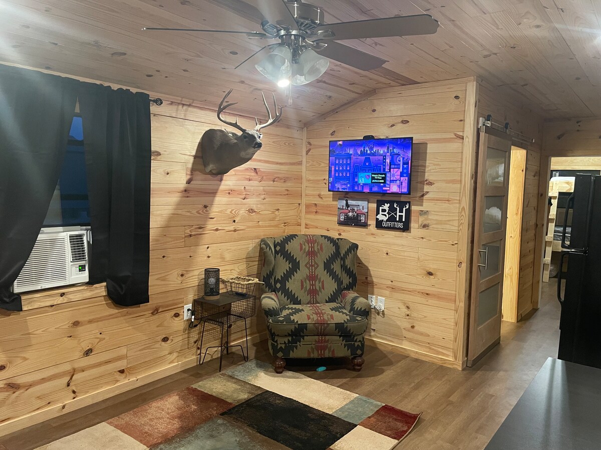 A cozy living area is defined by wooden walls and a neutral-toned rug. A comfortable patterned chair sits beside a small table. A mounted deer head and a flat-screen television are featured on the wall, while a ceiling fan provides ventilation.