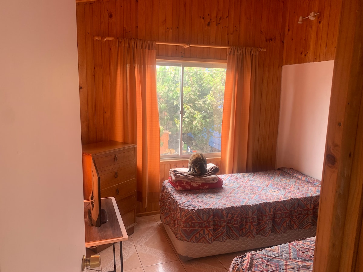 A well-lit bedroom is furnished with two separate beds, each covered in patterned blankets. A wooden dresser stands against one wall. Natural light enters through a window draped with orange curtains, illuminating the space and offering a view of greenery outside.