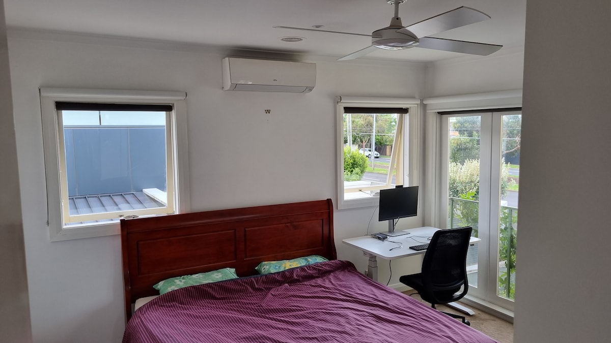 A spacious master bedroom features a king-size bed with a striped blanket. A desk with a computer chair is positioned by the window. Natural light enters through two windows, highlighting the room's neutral decor and air conditioning unit on the ceiling.