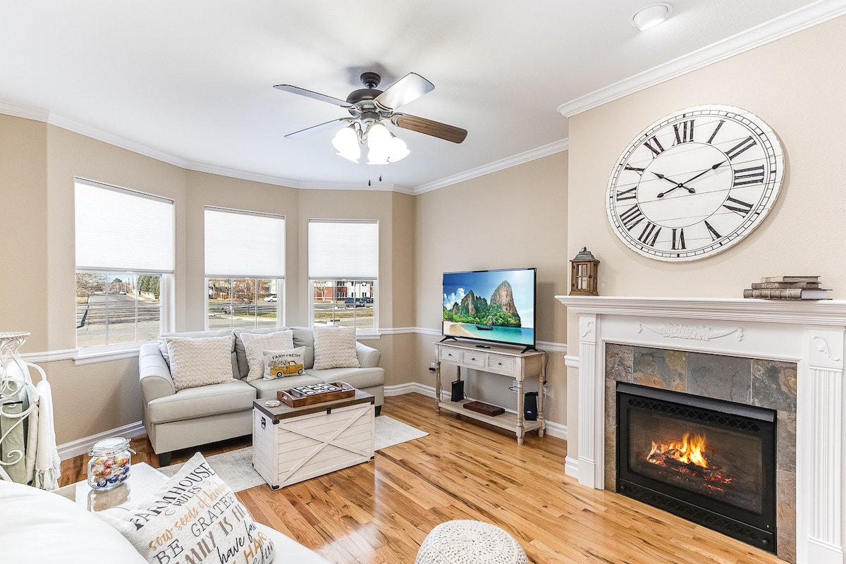 A spacious living room features a comfortable couch facing a wall-mounted television, with natural light from three windows. A decorative fireplace is situated under a large clock, and hardwood flooring adds warmth to the inviting atmosphere. A coffee table sits at the center of the seating area.