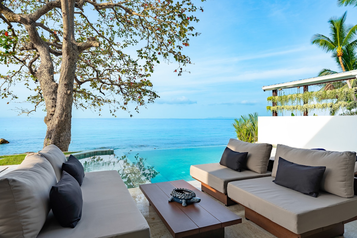 A spacious outdoor seating area is presented, featuring low-profile sofas with soft cushions, perfectly arranged to face the stunning ocean view. The inviting saltwater infinity pool is visible, blending seamlessly into the tranquil blue waters of the sea beyond.