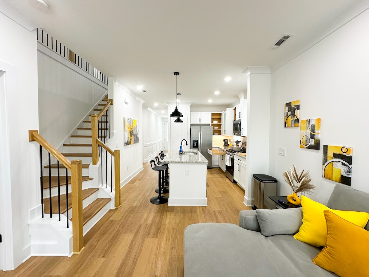 The open-plan living area features a modern kitchen with a large island, bar stools, and sleek appliances. A light wood staircase ascends to the upper level. The room is decorated with wall art in bright yellows and blacks, complemented by a spacious gray sofa adorned with vibrant yellow pillows.