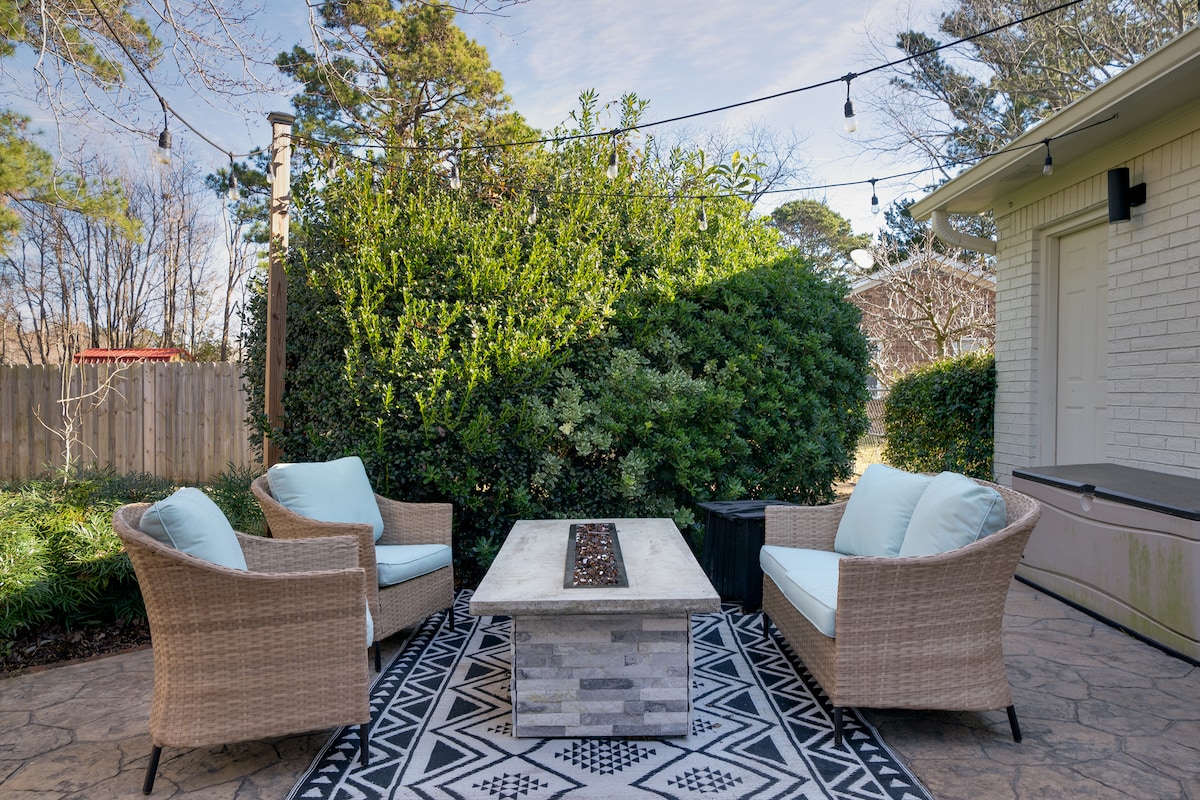 An outdoor seating area is showcased, featuring two comfortable chairs positioned around a modern fire table. Surrounding greenery provides privacy, and a patterned area rug adds texture to the space. Soft lighting is provided by overhead string lights, creating a pleasant ambiance.