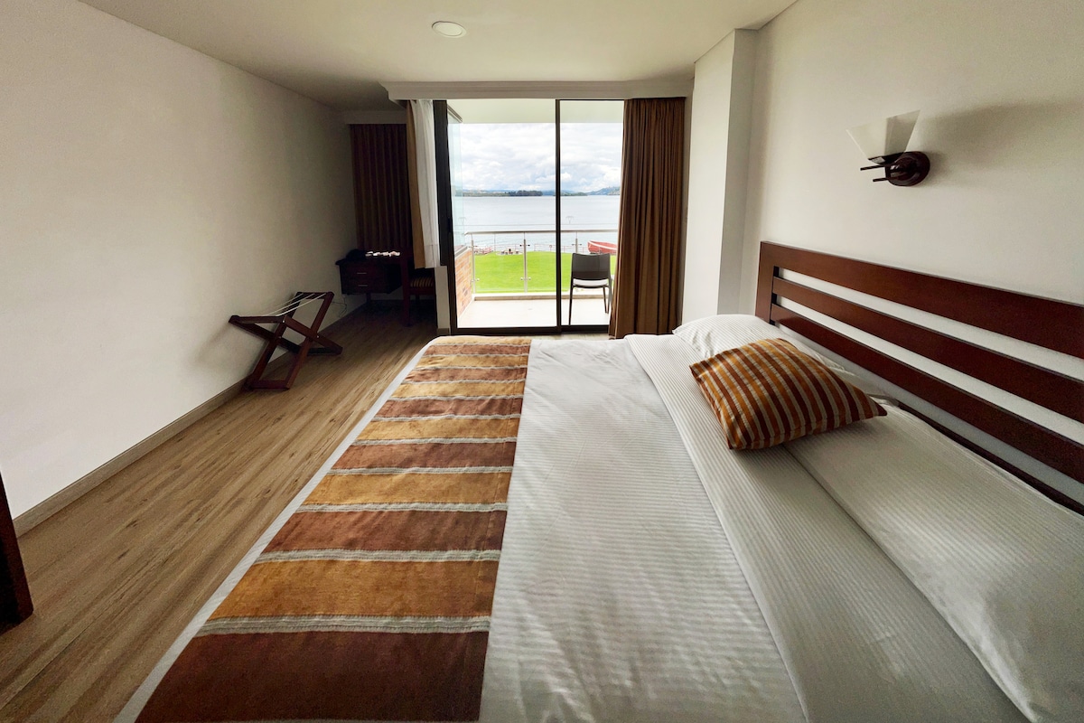 A bright sleeping area features a spacious bed with a striped accent pillow. Large glass doors open to a view of the lake, allowing natural light to fill the room. Simple furnishings include a wooden chair and a small table placed nearby.