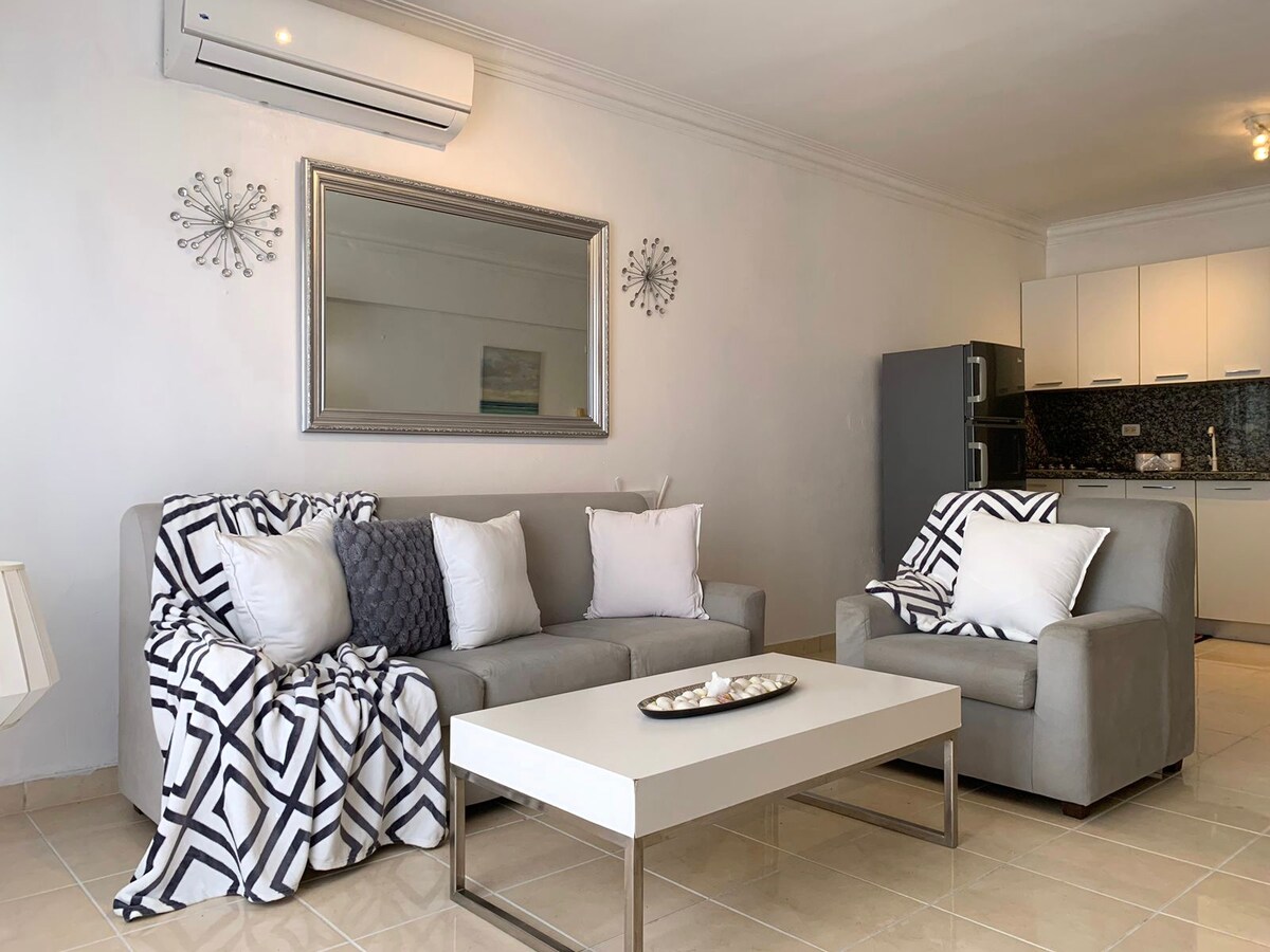 A comfortable living area features a light-colored sofa adorned with patterned throws and accent pillows. A contemporary coffee table is positioned in front, with a decorative tray displaying small shells. The space is complemented by a large mirror and modern kitchen appliances in the background.