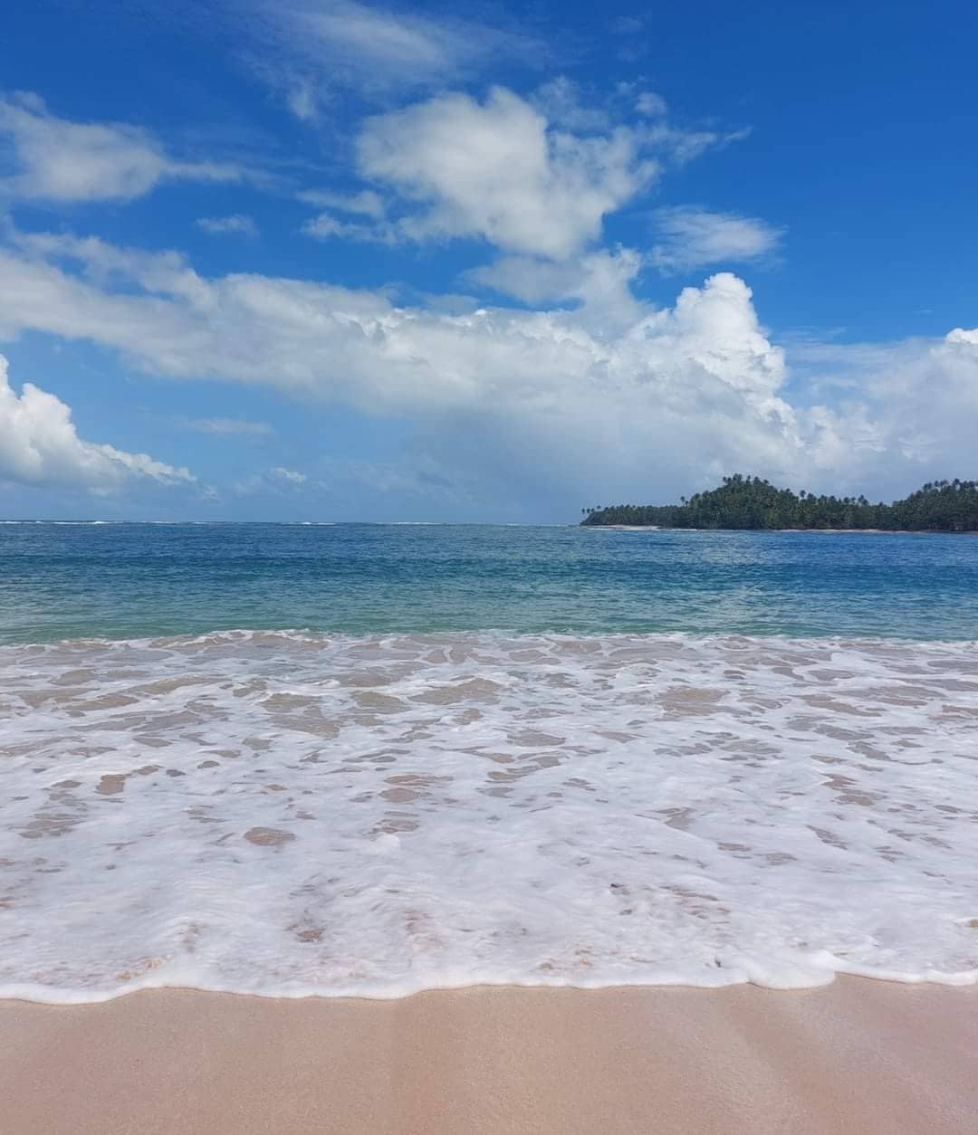 The view showcases a serene beach where gentle waves lap against soft, pale sand. Clear blue skies and scattered clouds create a tranquil backdrop, while distant greenery on the shoreline adds a touch of natural beauty to the scenic landscape.