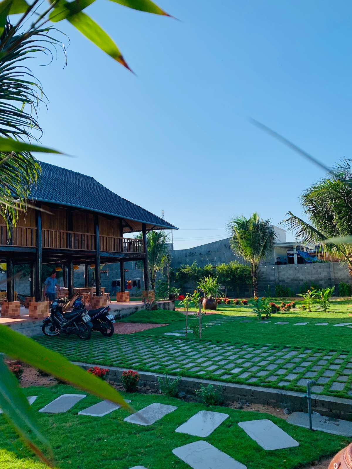 The exterior of a unique house is framed by lush greenery and palms. A spacious yard showcases a well-maintained garden with pathways and flowering plants. The two-story structure features wooden elements and is set against a clear blue sky.