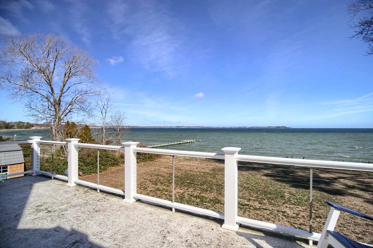 A spacious outdoor deck with a white railing offers a clear view of the Piankatank River. The rippling water reflects the blue sky, while a wooden pier extends into the distance. Nearby, grass and bare trees provide a natural backdrop.