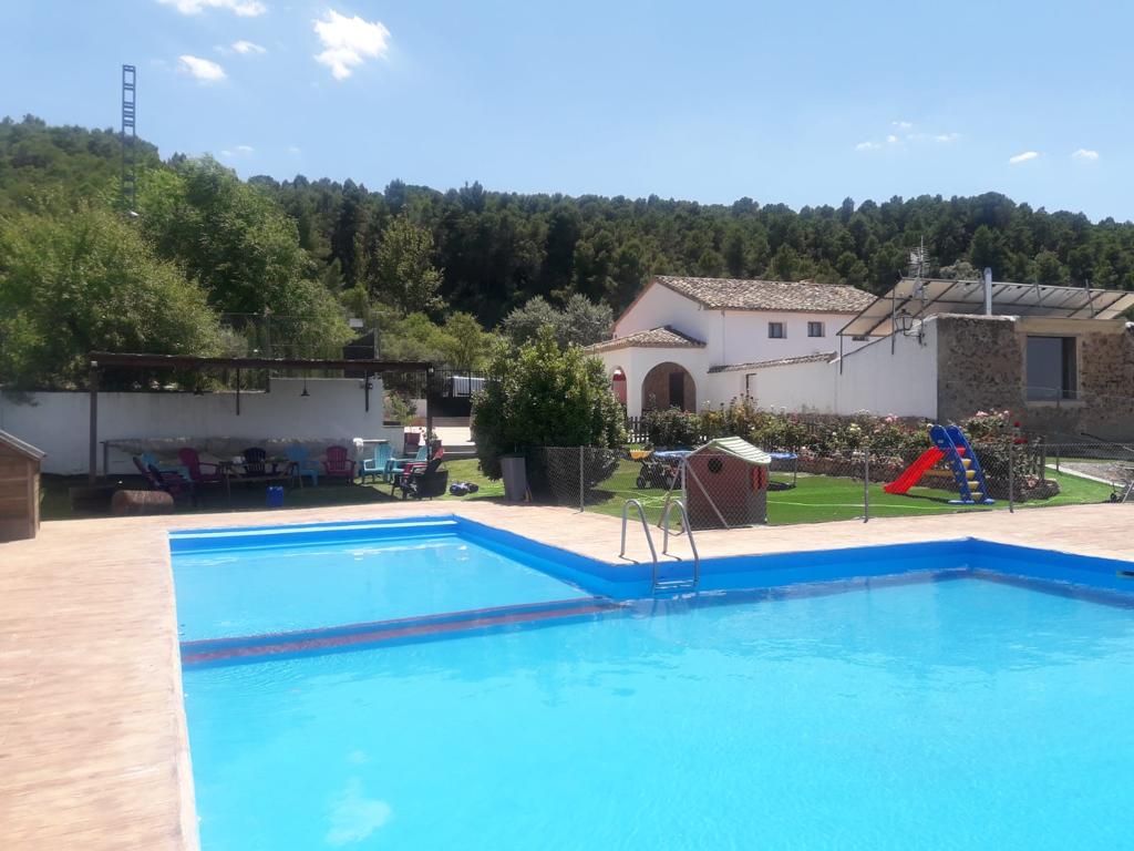 A clear swimming pool is set against a backdrop of lush greenery, with a landscaped area featuring a children’s play structure. A stone building with a traditional roof and outdoor seating areas can be seen, creating a welcoming outdoor space.