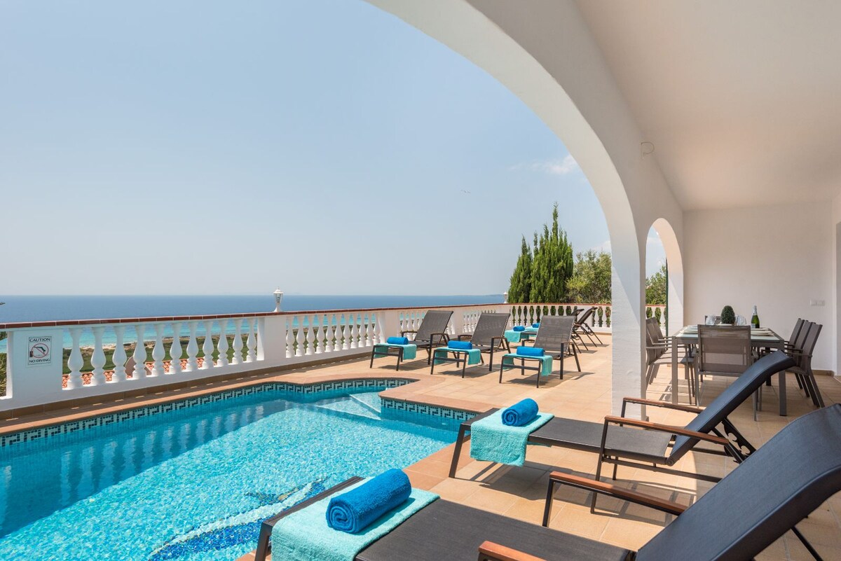 An outdoor area features a swimming pool surrounded by sun loungers, with blue towels neatly arranged on each. A dining table is positioned nearby, with the ocean visible beyond an elegant railing. Lush greenery is present in the background, complementing the tranquil setting.