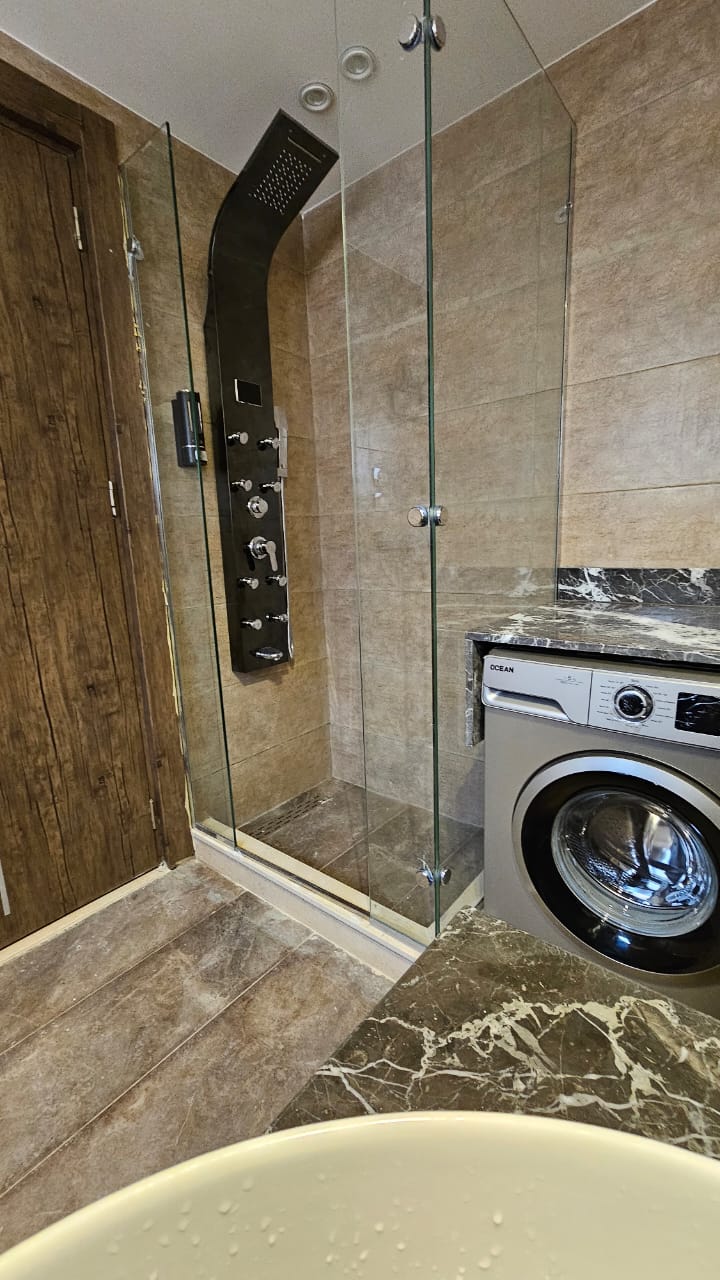 A modern bathroom features a glass-enclosed shower with multiple showerheads, accompanied by a sleek washing machine. Brown wood accents complement the tiled walls, and a marble countertop adds to the contemporary design.