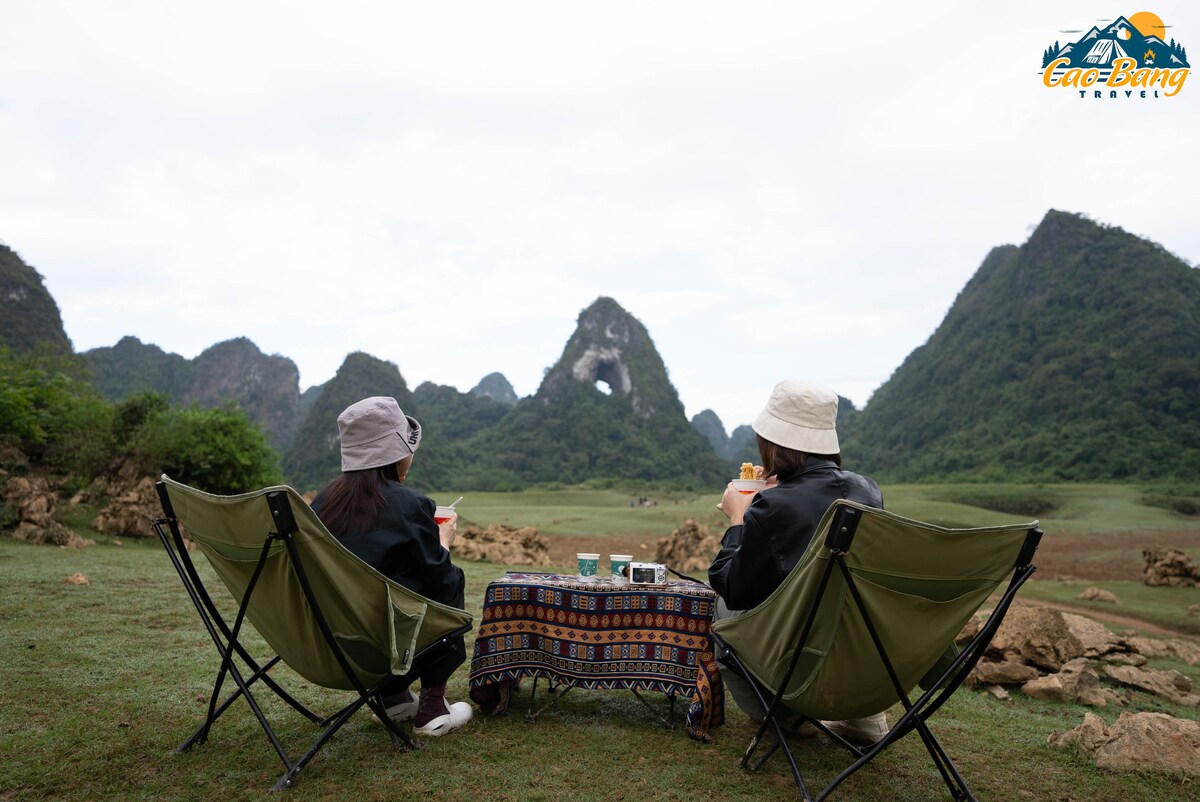 Two individuals are seated in green camping chairs, enjoying beverages while facing a view of mountains. The distinctive mountain features a prominent hole, with lush greenery surrounding the open area. A patterned blanket is spread over the ground, adding a cozy touch to the outdoor setting.