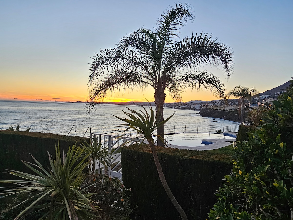 A serene view of the ocean is framed by palm trees and lush greenery. The horizon showcases a sunset with shades of orange and yellow, while the calm waters reflect the colors. A glimpse of a modern pool can be seen, contributing to the tranquil ambiance.