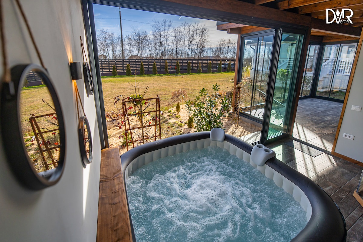 An elegant jacuzzi sits in the foreground, surrounded by windows that frame a scenic view of the green landscape outside. The bubbling water is inviting against the backdrop of a serene outdoor space, enhancing the atmosphere of relaxation.