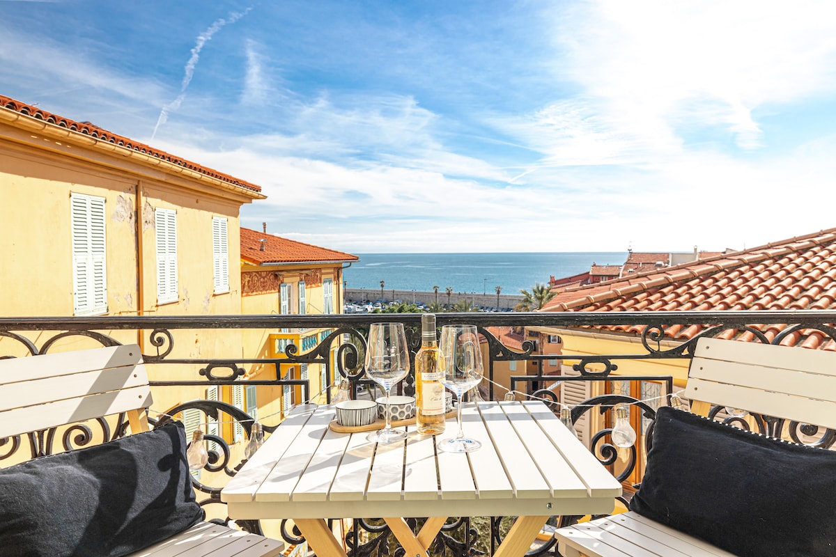 A balcony table set with two glasses and a bottle overlooks the sea, framed by a wrought-iron railing. The vibrant yellow buildings and red rooftops of the historic district are visible in the background under a clear blue sky.