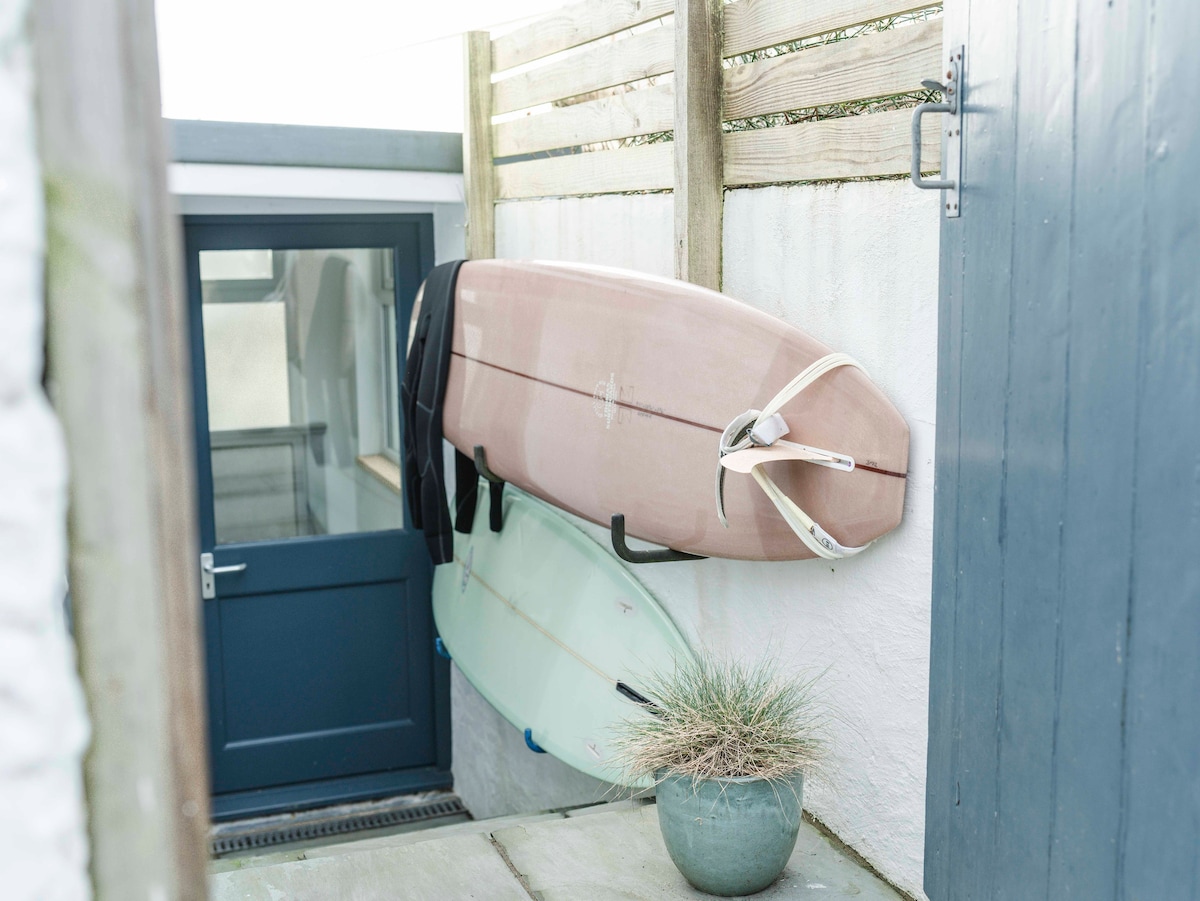 Two surfboards are mounted against a textured wall, showcasing a light-colored board and a pastel pink board. A door with a dark blue finish is visible to the left, while a minimalist plant arrangement sits at the base, enhancing the corner space.