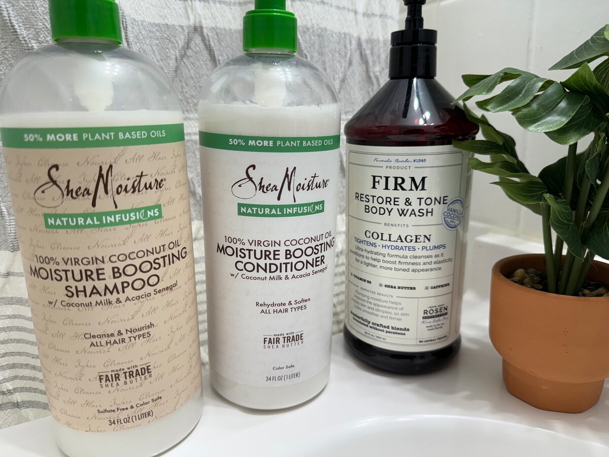 Three bath products are arranged on a bathroom counter. They include a shampoo, a conditioner, and a body wash, all labeled with their specific benefits. A small green plant adds a touch of natural decor, complementing the products.